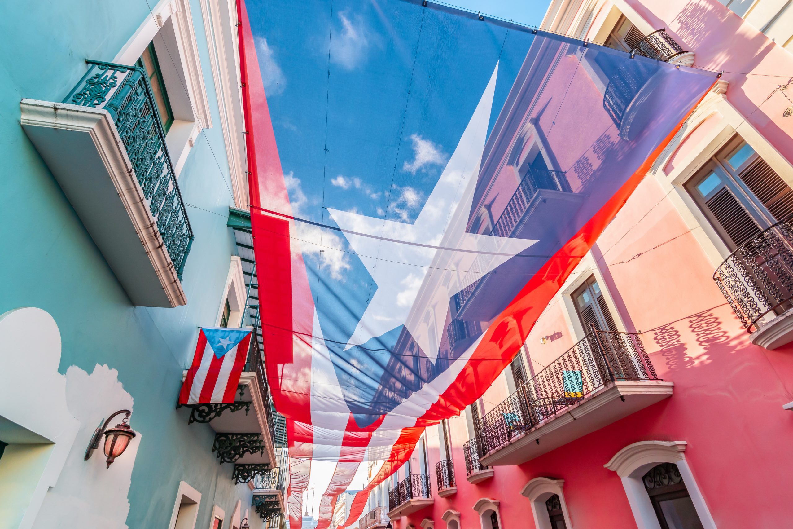Colorful image of city centre of San Juan with large Puerto Rican flag above the street. Blue and pink buildings in the street. Sunny day. Red and white stripes, white star and blue colored national flag of Puerto Rico.